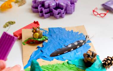 Child smearing colorful plasticine on cardboard and creating fairy tale card with cartoon animals, using nature details also, dry flowers, cone, leaves. Hobby. DIY
