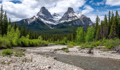 Twin mountain peaks reach towards cloudy skies, framed by a rocky riverbed and dense evergreen forest under a bright, sunny sky