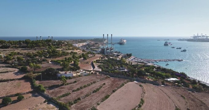 Malta: Aerial view capturing Delimara Power Station in Marsaxlokk, Malta, featuring the power plant, fuel storage tanks, and cargo ships in the Mediterranean Sea. Drone flight footage