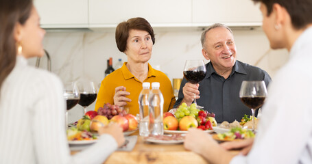 Elderly mother with surprised expression listening to young couple while sitting at cozy set table during family gathering