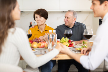 Puzzled elderly mother looking on with concern and surprise while adult son in love engaging in conversation with girlfriend at dinner table during family meal at home, young couple in love clasping