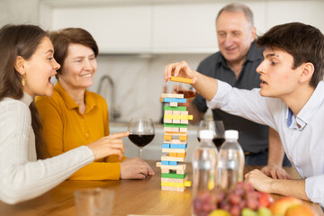 Young guy playing jenga with family in cozy home kitchen, trying to put brick without destroying...