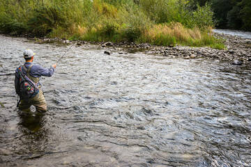 Fisherman casting fishing line while standing in a river, surrounded by lush greenery