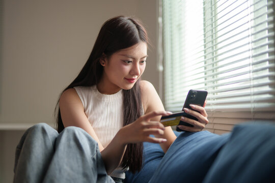 Happy customer making purchase in online store using mobile phone and paying with credit card sitting on sofa at home