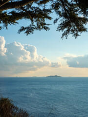 A view of the calm blue sea from a high cliff with distant islands on the horizon framed by pine tree branches under a partly cloudy sky a serene and beautiful seascape