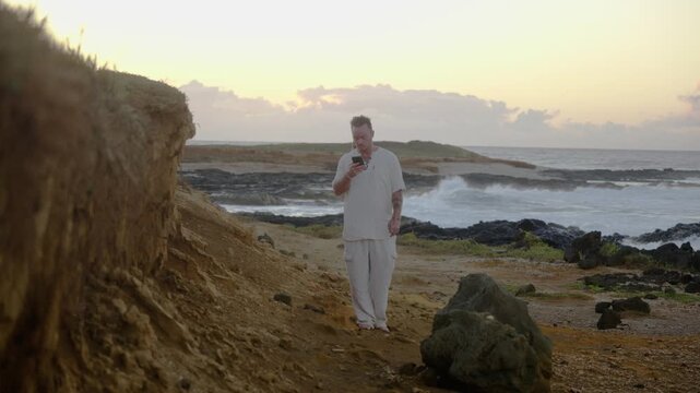 A man in white clothing walks toward the camera on a rugged coastal path, checking his smartphone as the ocean waves roll in under soft morning light.