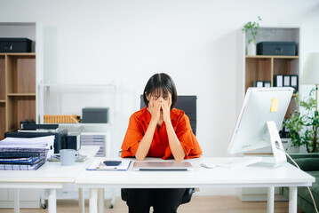 Asian woman overwhelmed at work, covering her face at a cluttered desk. Concept of stress, burnout,...