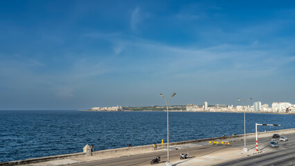 Havana Malecon. Cars, motorcycles on an asphalt road. People are walking along the embankment. Traffic light. Urban multi-storey houses in the distance. Ripples on the ocean surface. Blue sky, clouds