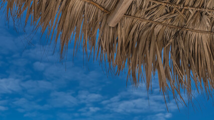 A straw umbrella on a background of blue sky and clouds. A fragment. Close-up. You can see the weaving, the brush of straw. Bottom-up view