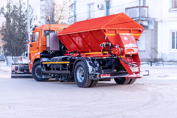 Orange municipal snow removal machine with sand and salt spreader. Road service truck cleaning street from snow. Winter utility transport for clear roads.