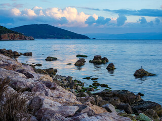 A beautiful coastal view of a rocky shoreline covered in green moss with a distant hilly island across the calm blue sea under a soft sky