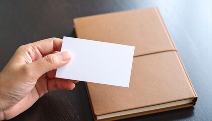 Obraz premium A person's hand holding a blank white business card over a brown journal, ideal for a professional branding mockup