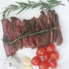 A steak on a white plate ready to eat, garnished with red tomatoes and rosemary for flavor.