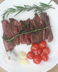 A steak on a white plate ready to eat, garnished with red tomatoes and rosemary for flavor.