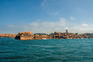 Scenic view of the Pink Granite Coast in Brittany, France, with a lighthouse on rocky shore, vivid blue sea, and clear sky. Natural coastal landscape with unique rock formations.