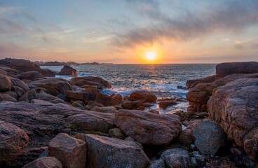 Dramatic sunset over the Pink Granite Coast in Brittany, France. Waves crashing on the rugged rocks, vibrant sky colors reflecting on the sea. Peaceful ocean landscape with natural stone formations.