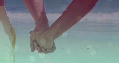 Holding hands of couple wearing light beach shorts, pink sleeveless top in shallow surf, soft flare