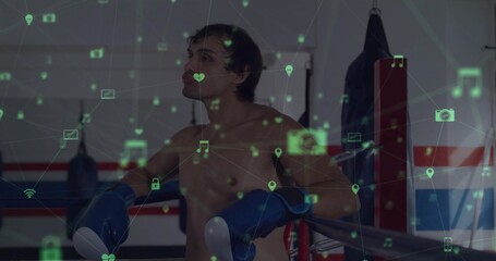 Leaning shirtless boxer wearing blue gloves on ropes by punching bags at gym, showing digital icons