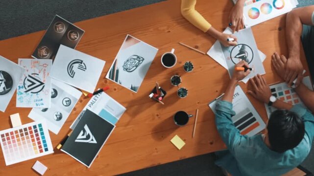 Top view of designer writing and designing logo graphic at meeting. Closeup of business man working on colored palettes and writing idea on sticky notes on table with equipment and paper. Symposium.