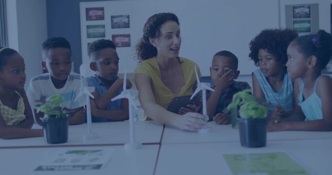Gesturing teacher in yellow blouse explaining wind turbine models at classroom table, with tablet