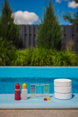 Chlorine tablets and a pool water test kit with chemical dropper bottles and color scale are arranged on the poolside in sunlight, used for pool maintenance.