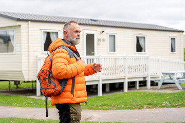 Middle-aged man in bright orange jacket and backpack walking near a mobile home at a campsite...