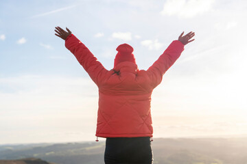 Person wearing a red jacket raises arms in celebration on a mountain during sunset with a vibrant sky and rolling hills in the background