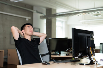 Fototapeta premium A young man leans back in his chair with hands behind his head, gazing upward in thought. He is daydreaming in the office.