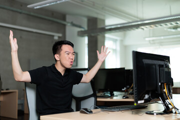 A young man throws his hands up in excitement while looking at a computer screen in a bright classroom. His energetic expression conveys a sense of triumph and joy.