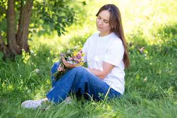 Beautiful woman with long hair sitting on green lawn holding a bouquet of wildflowers outdoors