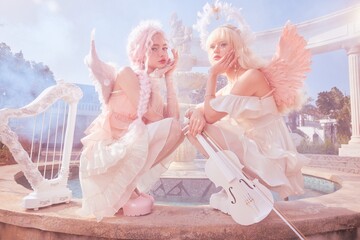 Two women in pastel outfits with wings posing gracefully by a fountain.