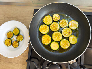 Fried zucchini slices in pan with oil and served on white plate, top view, homemade cooking process of healthy vegetarian or vegan meal.