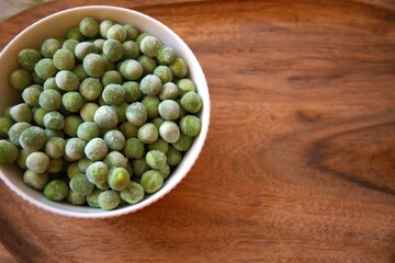 Fresh frozen green peas served in a white bowl on a rustic wooden table, close-up with copy space.