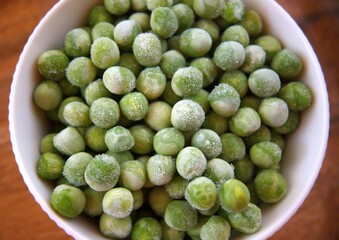 Fresh frozen green peas served in a white bowl on a rustic wooden table, close-up with copy space.