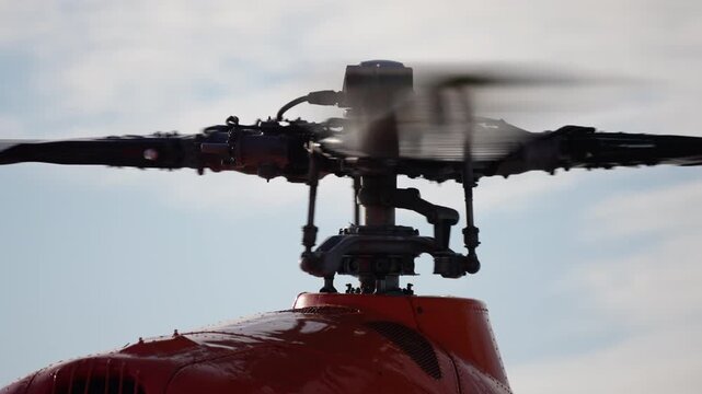 Close-up view of a red helicopter main rotor assembly in motion and coming to rest, highlighting the intricate hub, mast, and blade mechanisms against a bright sky.