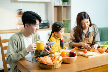 Asian family enjoys time together in kitchen as daughter uses tablet and mother prepares fresh food. Concept of parenting, home learning, and healthy lifestyle.