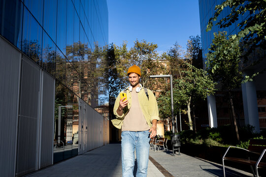 Stylish student walking and using smartphone between modern buildings - Powered by Adobe