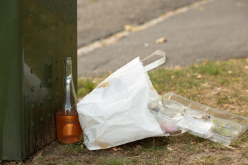 Paper bag, glass bottle, and plastic container with food remnants lying next to a public trash bin.