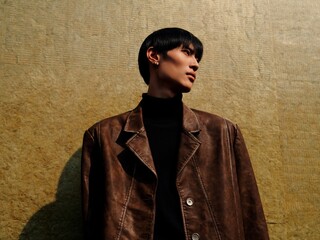 Stylish young man in brown leather jacket posing against a textured backdrop.