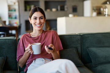 Portrait of a young latina woman having fun holding a remote control and watching tv at home