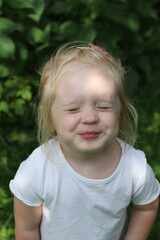 A blonde child in a white T-shirt stands in front of green leaves. The little girl squints from the sun.