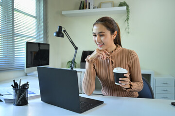 Smiling woman working on laptop at desk while holding a cup of coffee
