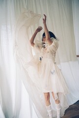 Teen girl in a white dress playfully dancing with flowing fabric indoors.