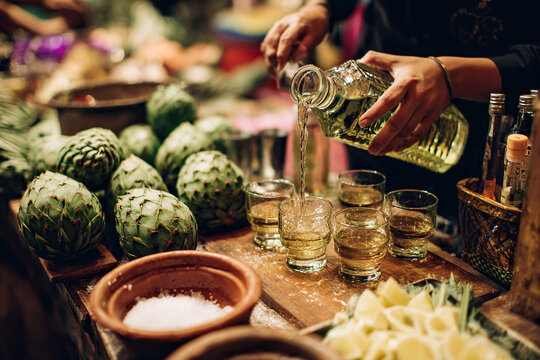 A bartender pours tequila into a shot glass on a bar counter decorated with agave, lime and salt. - Powered by Adobe