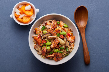 Salmon fried rice with mushrooms, red peppers and edamame, served with side dishes and a wooden spoon.