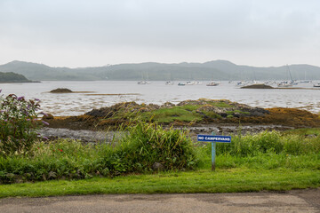 A 'No Campervans' sign is pegged into the ground at a lay-by overlooking Loch nan Ceall in the Scottish Highlands, with a view of the coast and bay seen behind. 