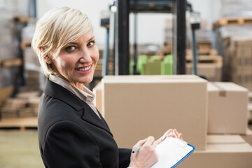 Middle-aged woman wearing blazer standing in warehouse holding clipboard near forklift, copy space
