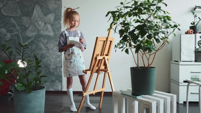 Little girl painting on easel with bright palette and focused face. Concept of early creativity, artistic expression, and talent development in childhood. Indoor natural scene