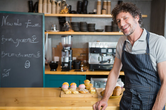 Male barista wearing apron leaning on cafe counter showing pastries, chalkboard menu, copy space - Powered by Adobe