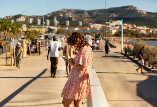 Jeune femme tapant sur son smartphone, vue de profil sur un trottoir le long de la mer, portant une robe d'&eacute;t&eacute; fleurie, adoss&eacute;e &agrave; une rambarde, promeneurs, ville et colline en arri&egrave;re-plan, Marseille
